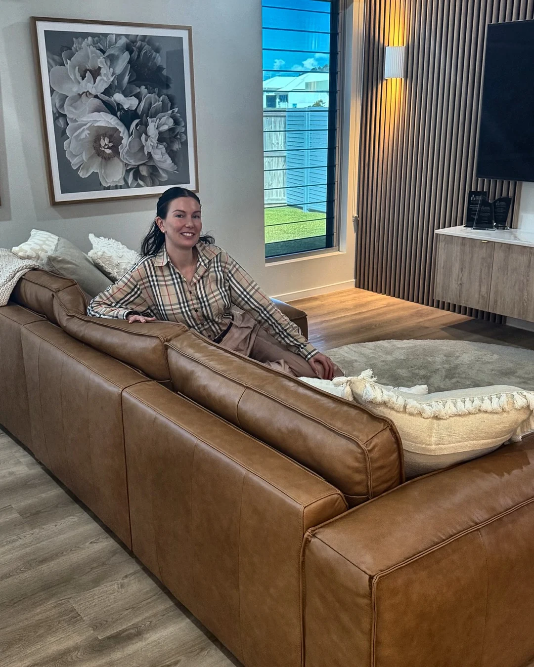 Melanie Laas smiling on a tan leather sofa in a living room with a timber slat wall