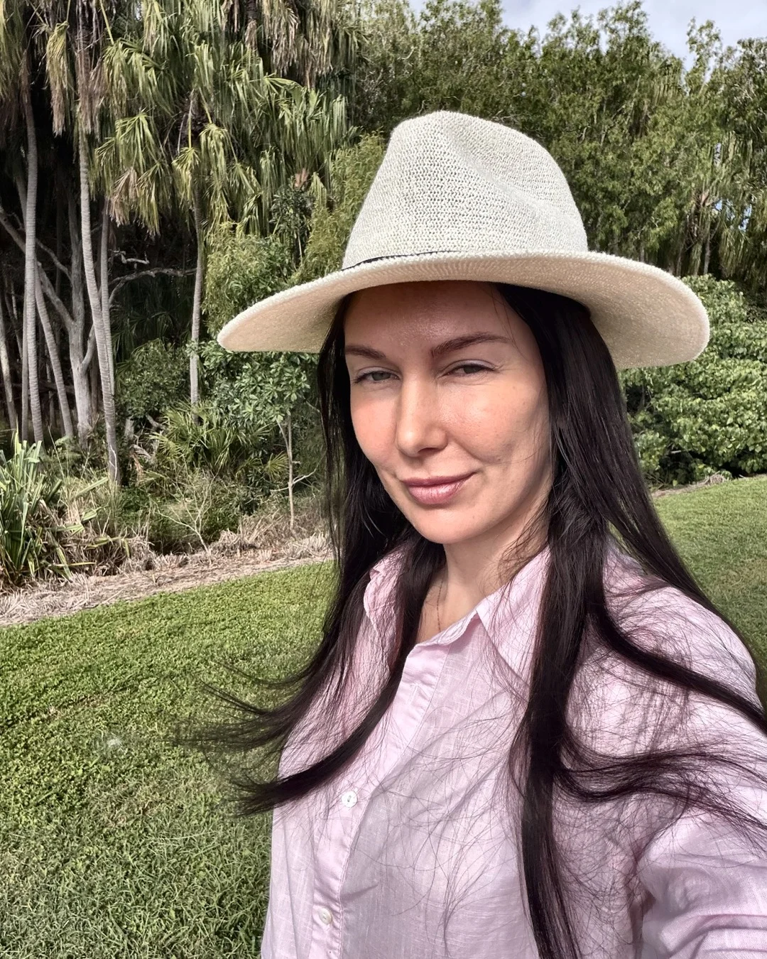 Melanie Laas in a cream hat and pink shirt, taking a selfie outdoors in front of palm trees