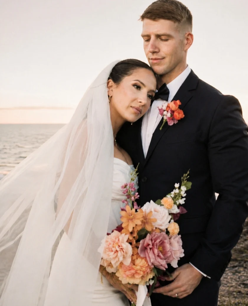 Bride resting her head on the groom's shoulder at a coastal wedding at sunset, holding a peach and pink bouquet