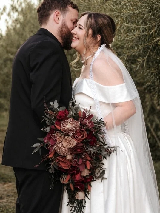 Groom kissing the bride's cheek in a garden, the bride holding a deep red and copper bouquet