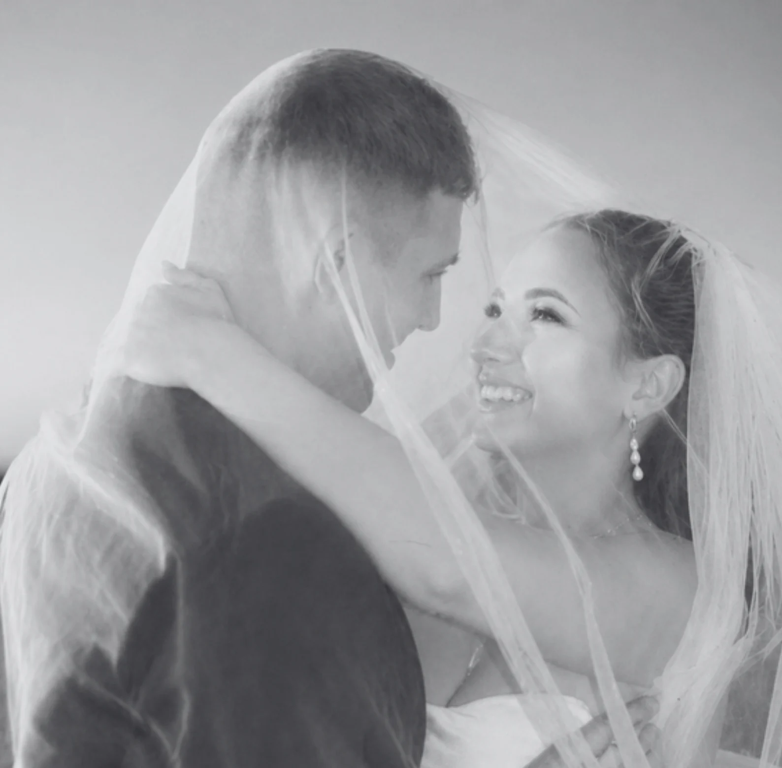 Bride and groom beneath a sheer veil, laughing together in a black and white candid wedding photograph by EMME Weddings