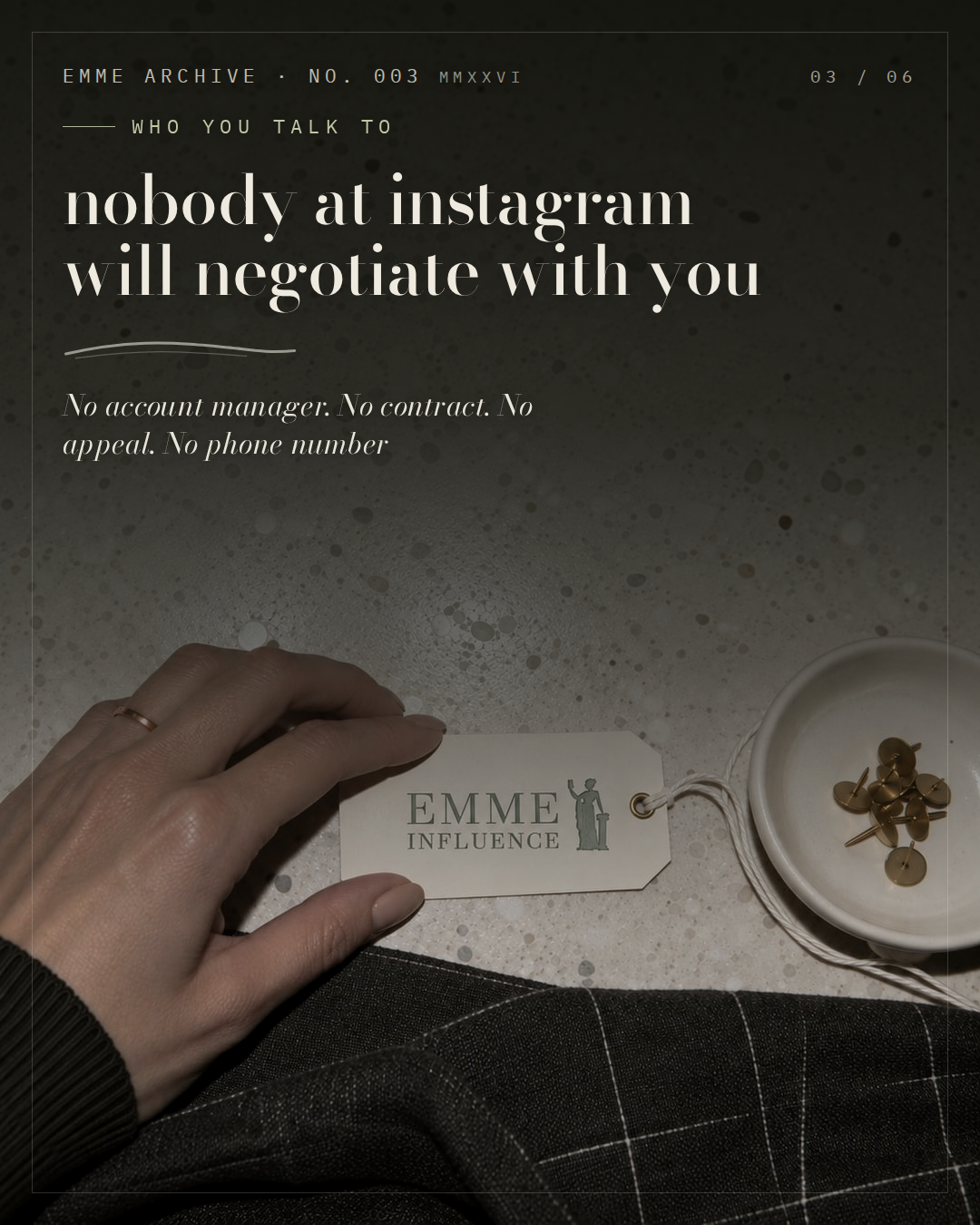 A hand pinning a cream archive tag printed with the EMME Influence mark, beside brass pins in a white ceramic dish